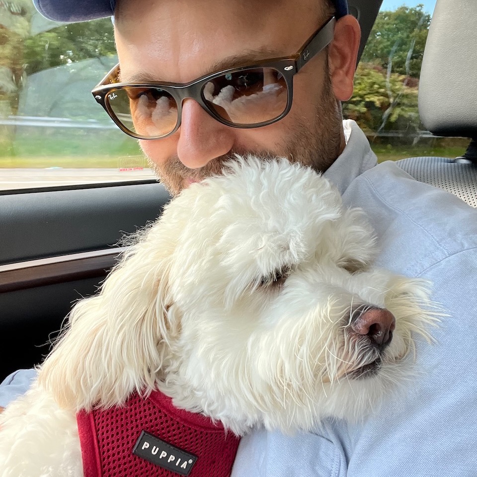 Chris, a white male with a short beard, wearing a light blue oxford shirt and Ray-Ban sunglasses. He has a small, fluffy white dog on his lap.